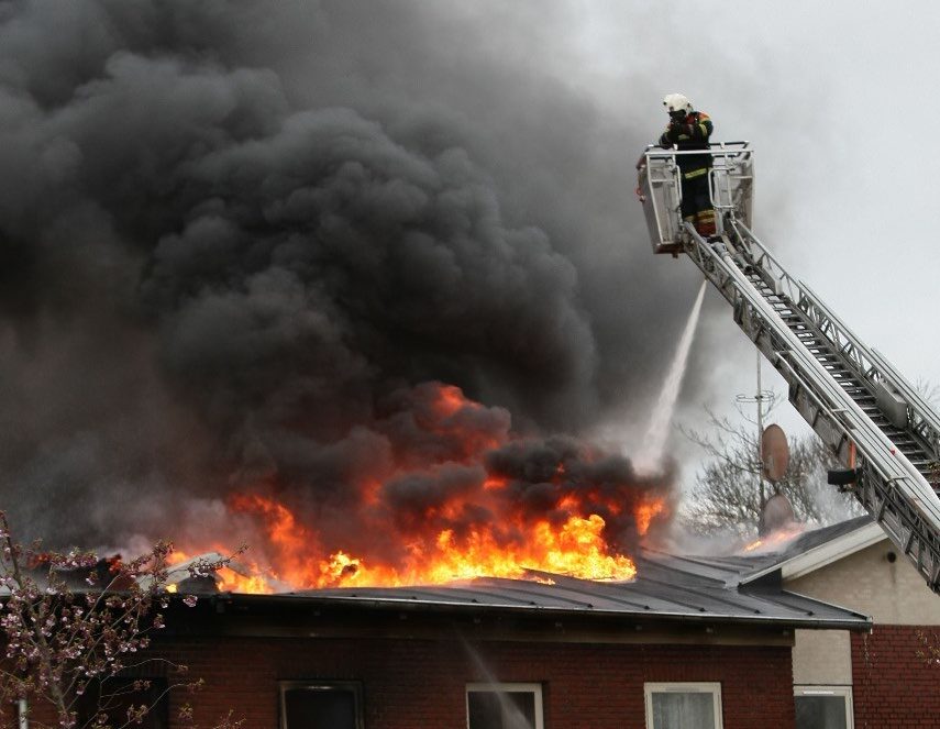 A fireman on an aerial ladder putting out a fire on the roof of a house with a fire truck hose.